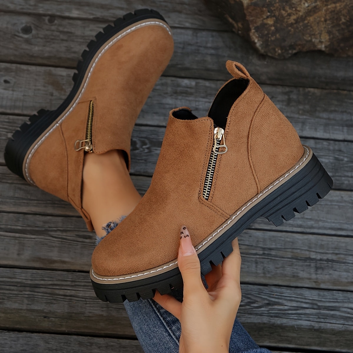 Brown ankle boot held above a wooden surface beside its pair, showing the side zip, pull tab, and lugged sole.
