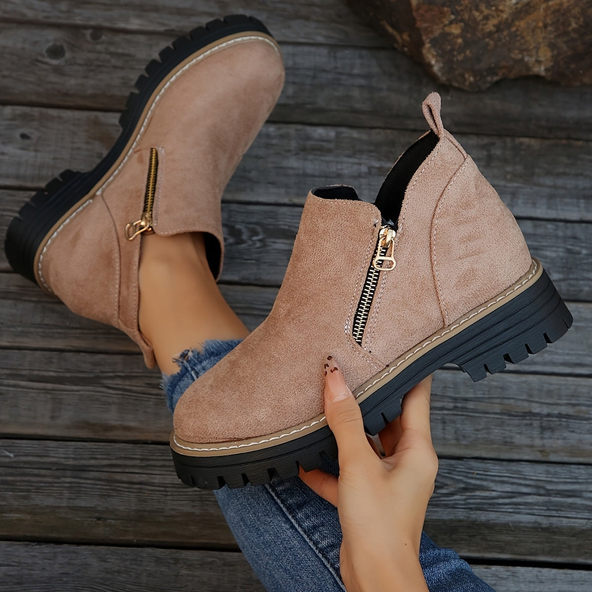 Blush ankle boot held in hand with its matching boot behind it, emphasizing the side zip, pull tab, and sole tread.
