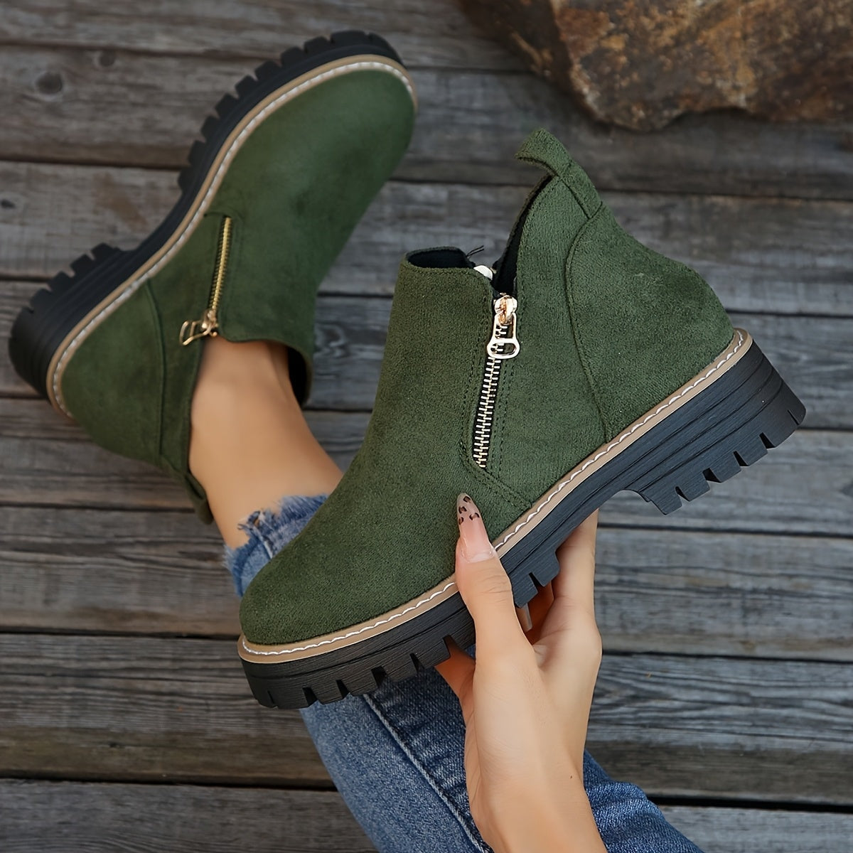 Green ankle boot held in hand with the matching boot in the background, highlighting the side zip and deep tread.