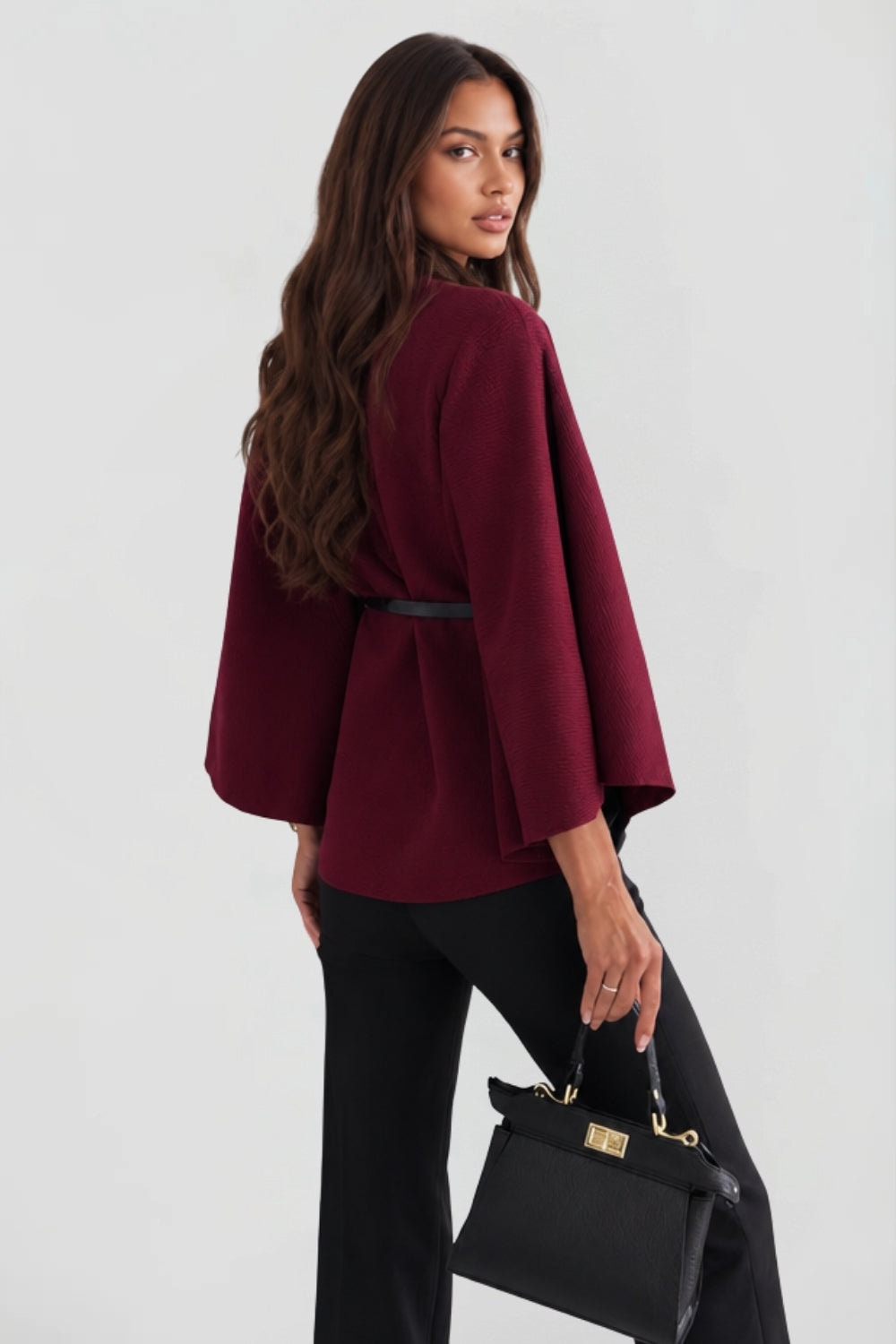 Back three-quarter view of the burgundy cape jacket, showing the belt across the back and flowing cape sleeves.