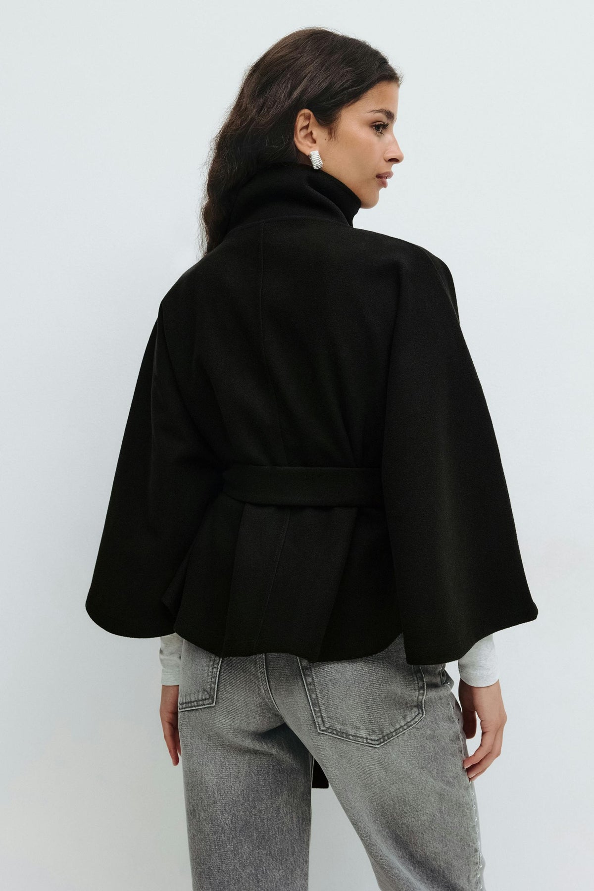 Back view of the black cape jacket worn over jeans, emphasizing its short drape, wide sleeves, and waist belt.