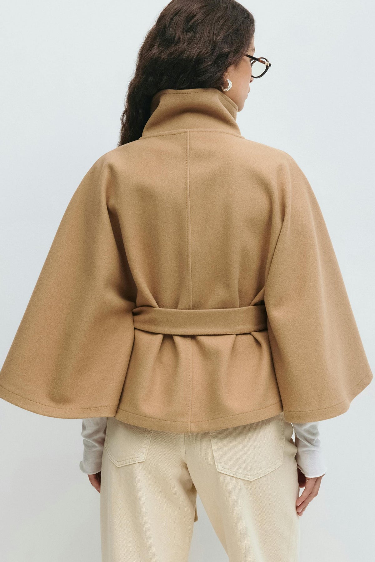 Back view of the camel cape jacket showing the center seam, broad sleeves, and belt wrapped around the waist.