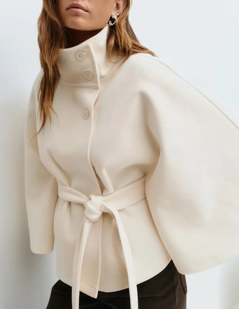Ivory cape coat shown at an angle with a raised buttoned collar, wide sleeve, and tied waist over dark trousers.