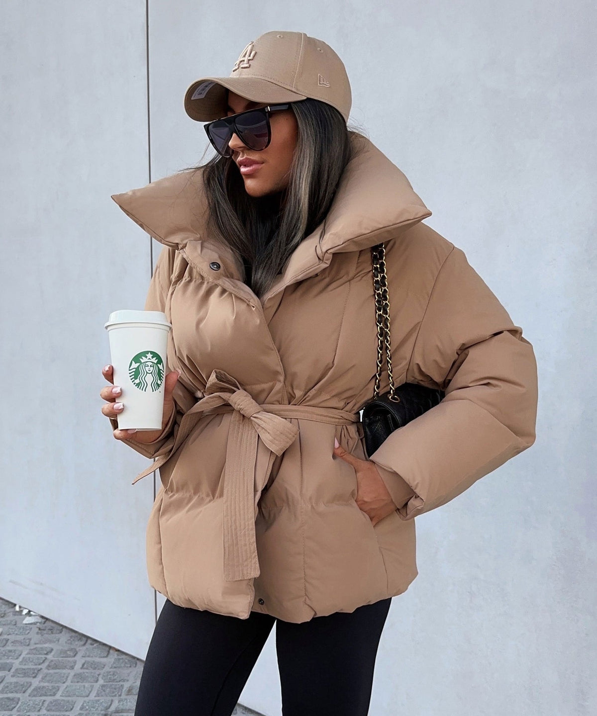 Taupe belted puffer jacket with a dramatic oversized stand collar, front zip closure, and hands placed in the side pockets.