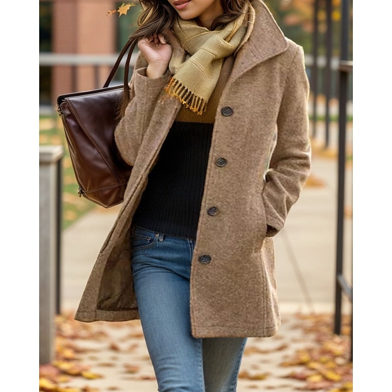Tan button-front coat with a notched collar shown open over a dark sweater and jeans on a leaf-covered sidewalk.