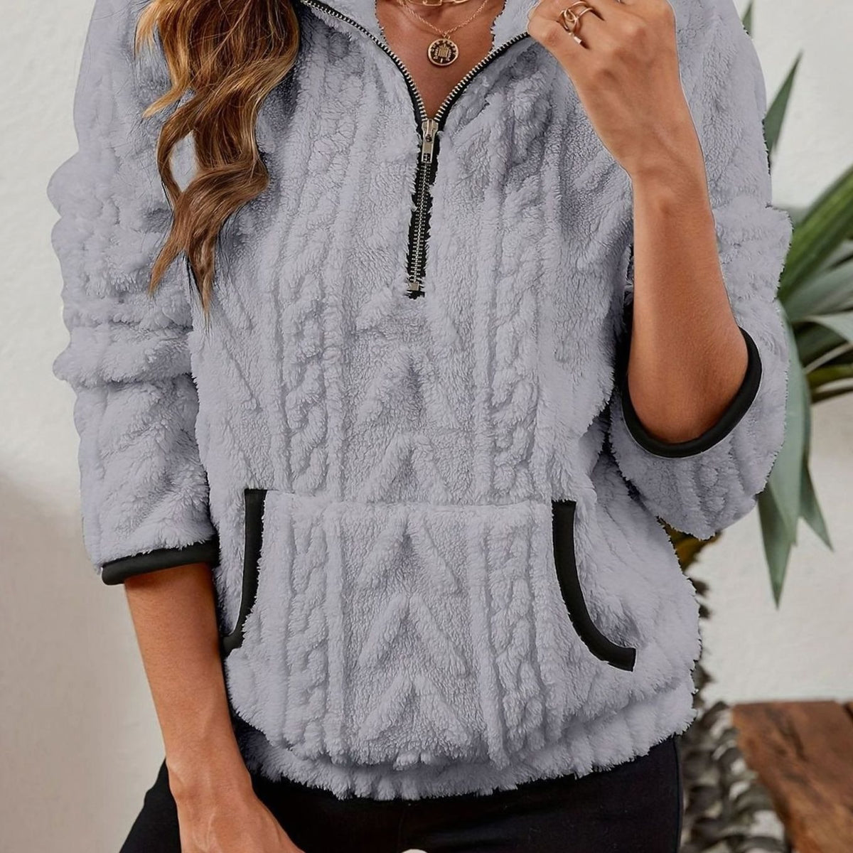 Close front view of the pullover in light gray, showing its raised cable texture, quarter zip, stand collar, and dark-trimmed kangaroo pocket.