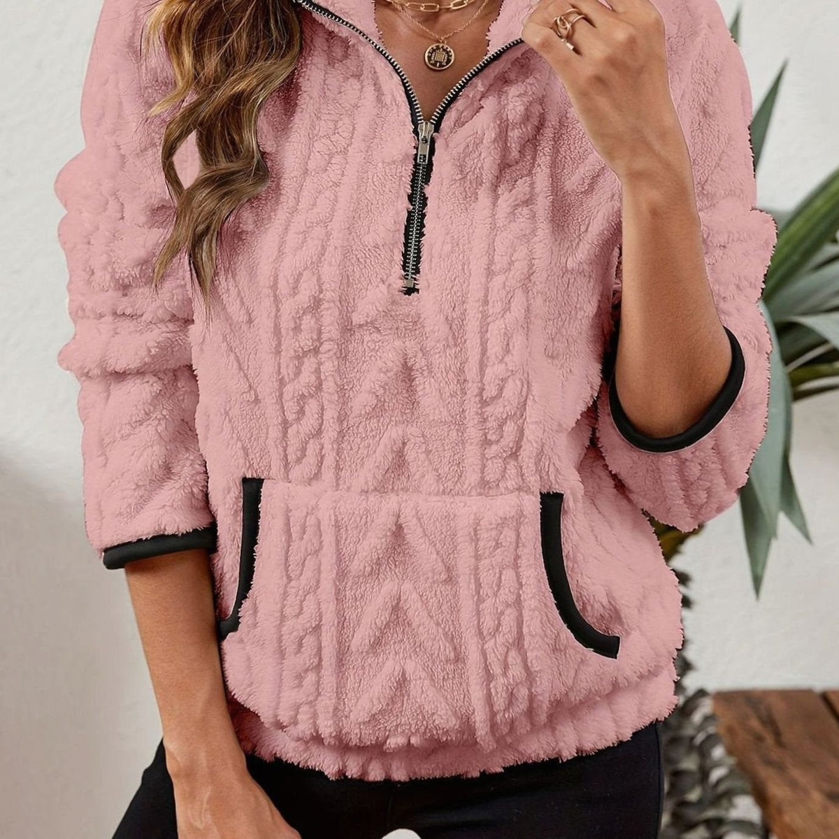Close front view of the pullover in pale pink with a raised cable pattern, half-length zipper, kangaroo pocket, and contrasting trim.