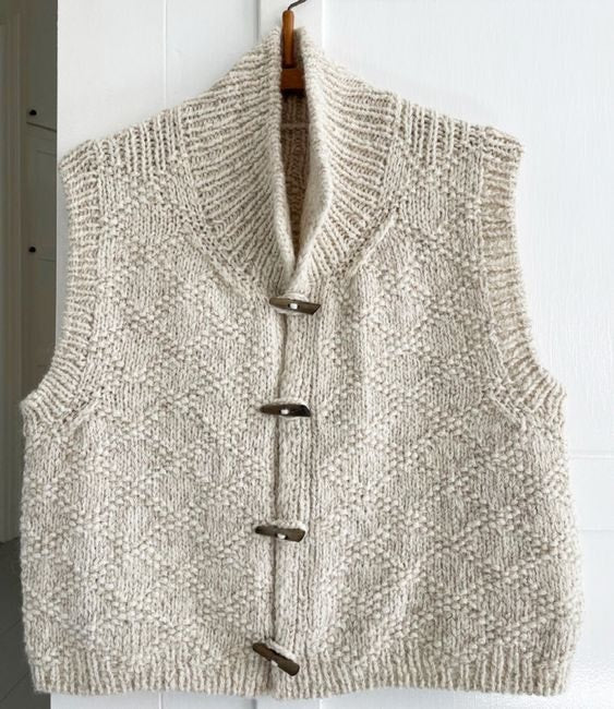 Full front view of the cream vest hanging on a hanger, showing its sleeveless shape, four toggles, ribbed hem, and textured knit pattern.