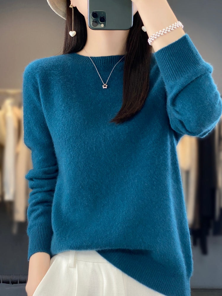 Deep teal crew neck sweater with a relaxed shape, long sleeves, and folded ribbed cuffs over light trousers.