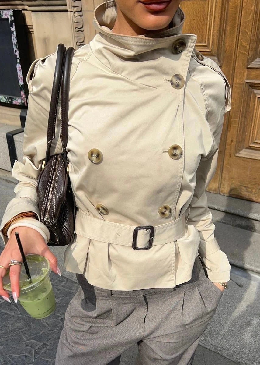 Close-up of the beige cropped trench coat showing its double-breasted button arrangement, raised collar, and rectangular belt buckle.