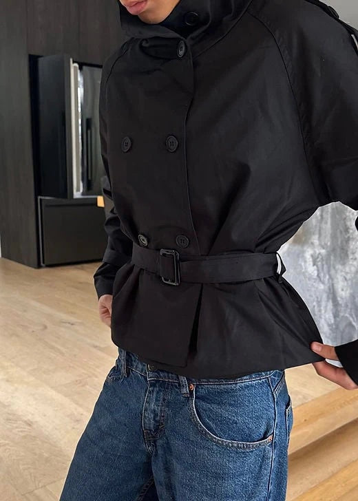 Close-up of the black version of the cropped double-breasted trench coat with a belted waist and raised collar.