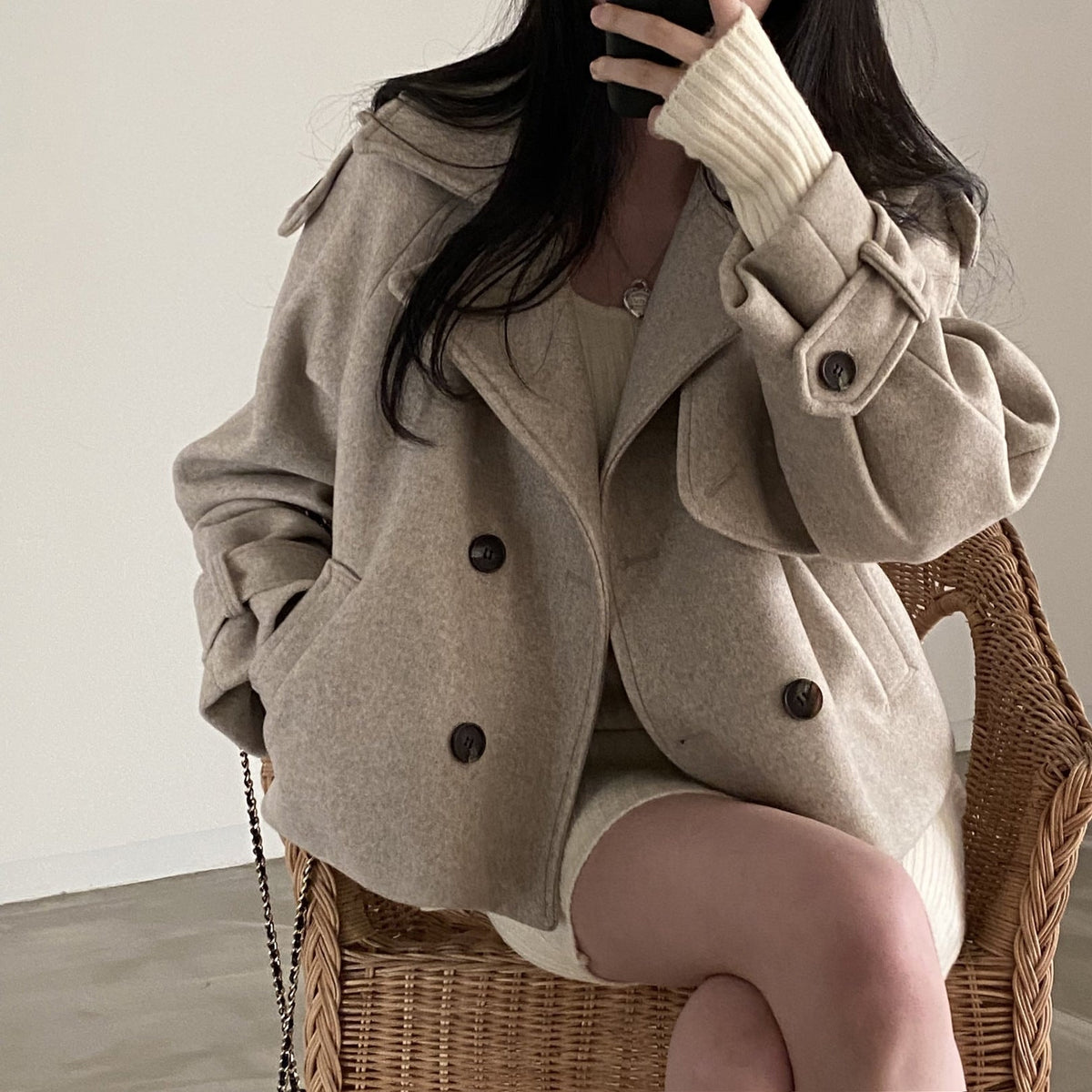 Seated front view of the taupe cropped peacoat styled over a light outfit, highlighting the double-breasted button arrangement, wide collar, side pockets, and gathered tabbed sleeves.