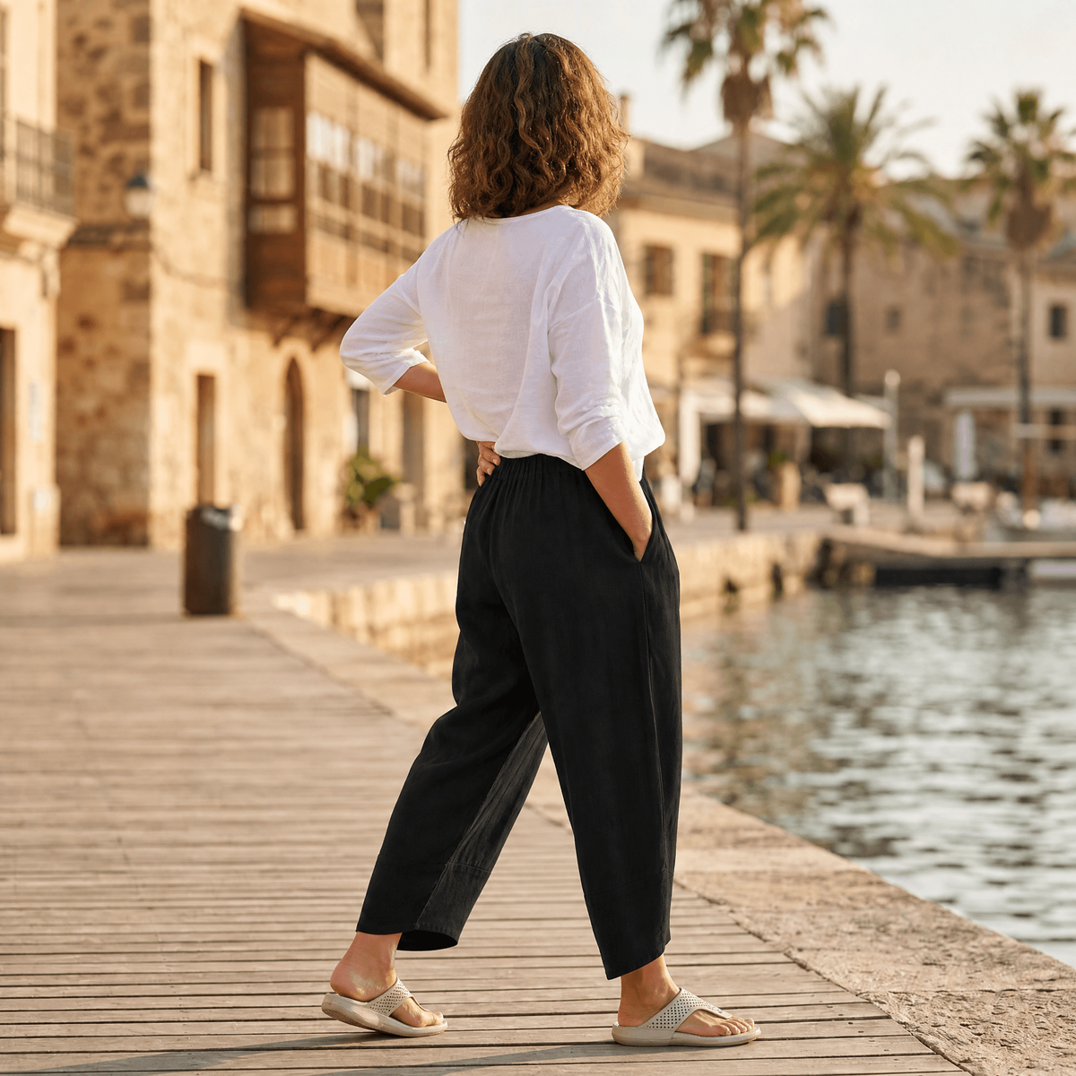 Rear three-quarter view of black cropped wide-leg pants with an elastic waist and side pockets on a waterfront boardwalk.