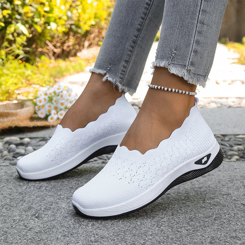 White knit slip-on walking shoes worn on both feet, featuring scalloped openings and contrasting dark soles.