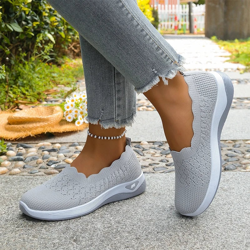 Gray knit slip-on shoes shown from opposite angles, including one raised shoe and one resting on the ground.