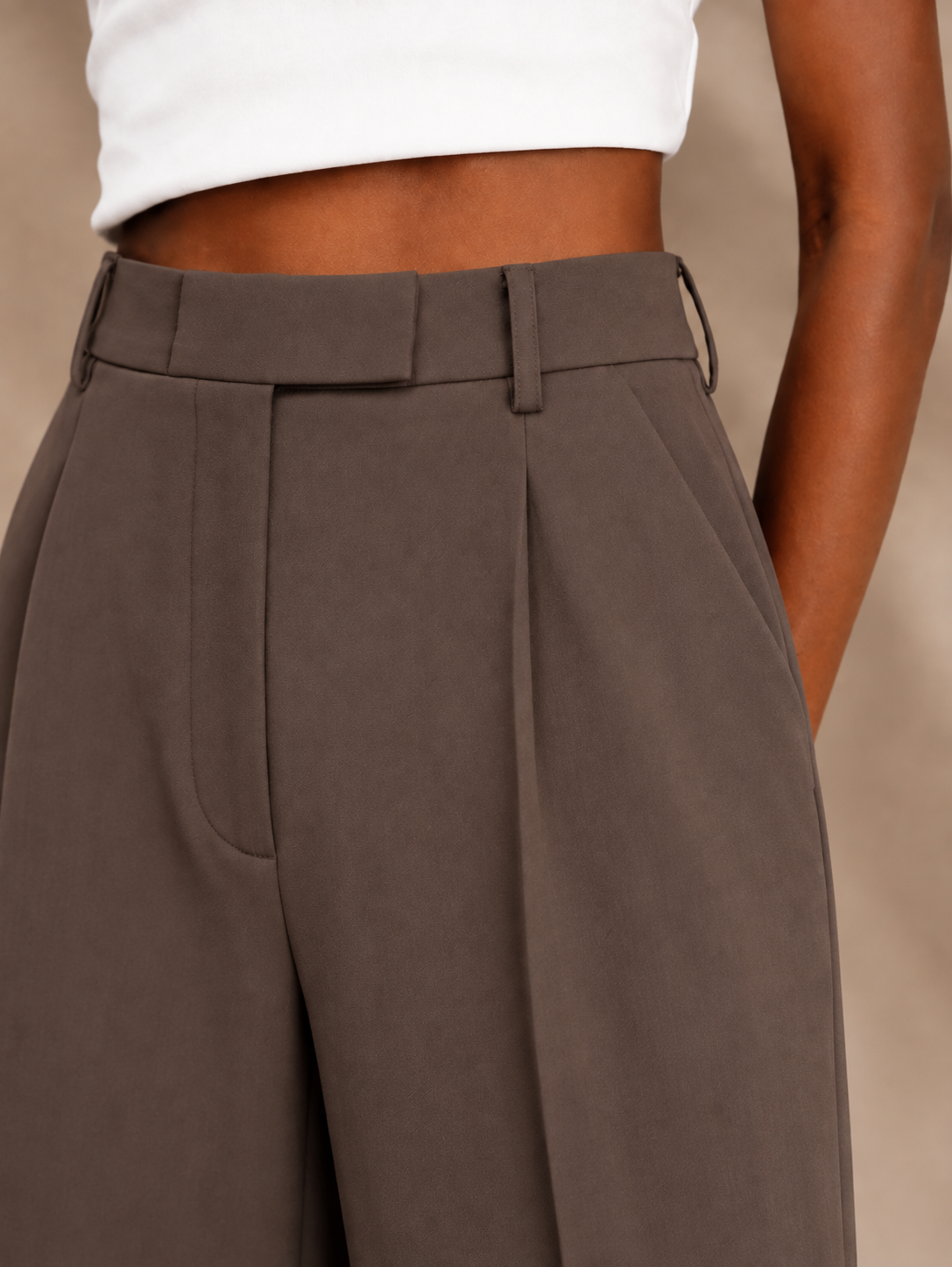 Close-up of brown trousers emphasizing the high waistband, front pleats, belt loops, and pocket opening.