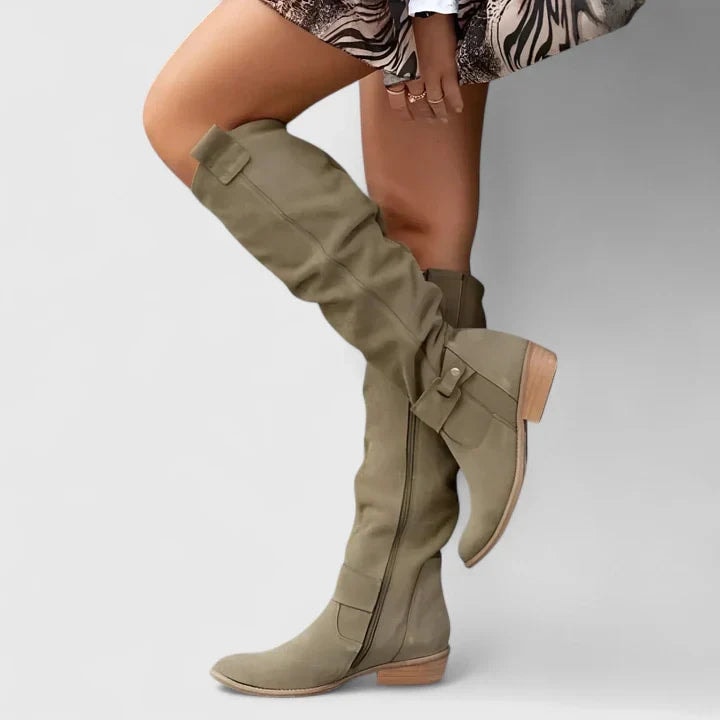 Olive knee-high boots with rounded toes, ankle straps, buckle accents, side zips, and low block heels shown crossed while worn with a patterned skirt.