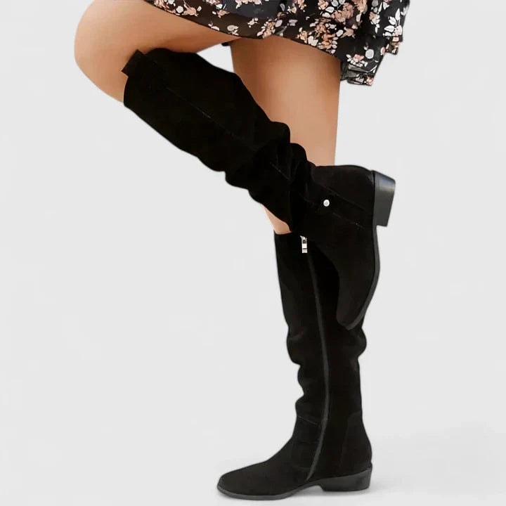 Black knee-high boot shown in side profile, emphasizing its tall ruched shaft, side zip, ankle strap with a button accent, and low heel.