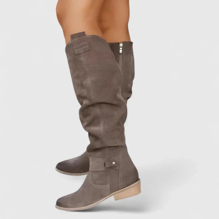 Taupe knee-high boots shown from the side with relaxed ruched shafts, visible side zips, ankle straps, buckle accents, rounded toes, and low heels.