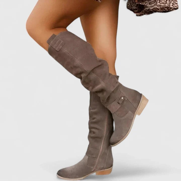 Taupe knee-high boots shown crossed in a raised pose, highlighting the slouchy shafts, ankle buckle details, rounded toes, side zip, and low block heels.