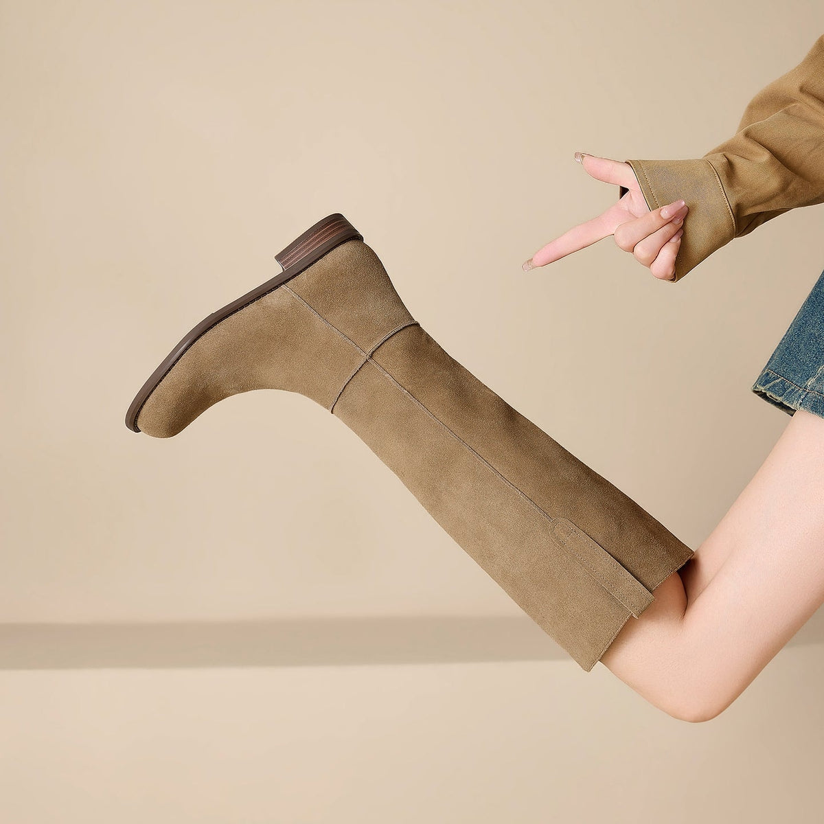 Close side view of a tan knee-high boot lifted horizontally, clearly showing its textured finish, seam, rounded toe, and stacked heel.