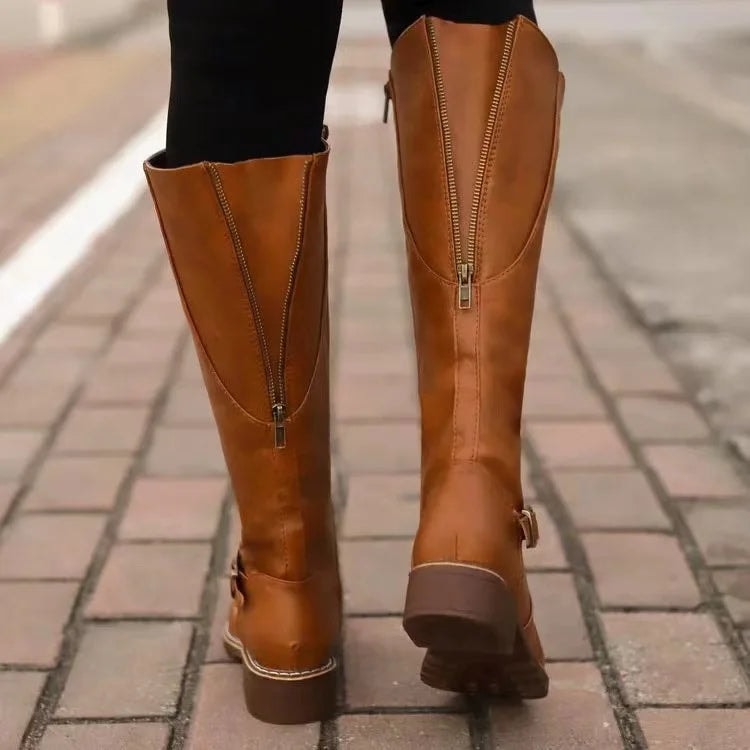 Tan knee-high riding boots viewed from the back while walking, highlighting the full-length rear zippers and block heels.