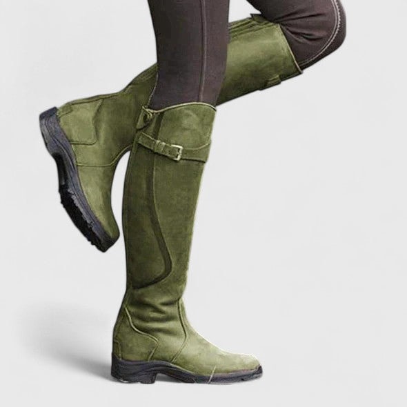 Green knee-high riding boots worn with dark leggings, highlighting the adjustable buckle strap, tall shaft, rounded toe, and textured outsole.