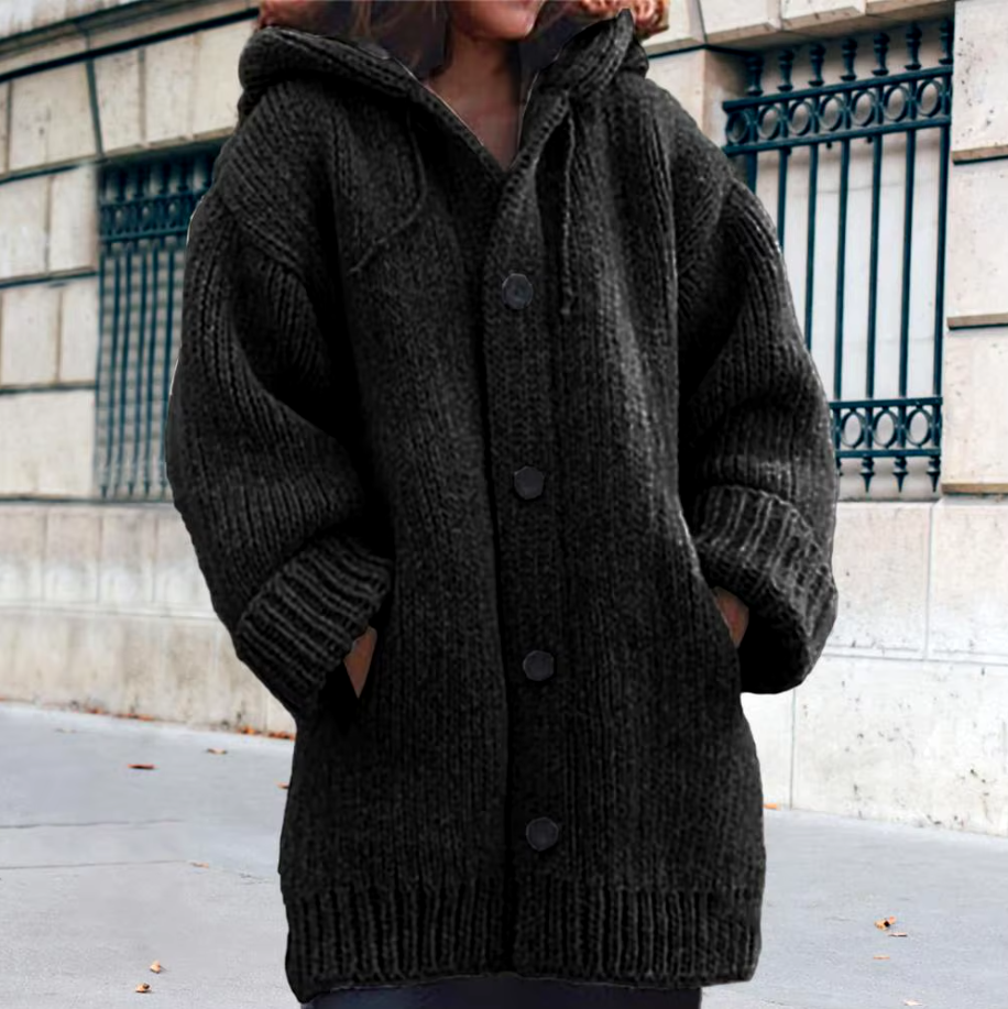 Black hooded chunky knit cardigan shown fastened with front buttons and hands placed in its two patch pockets.
