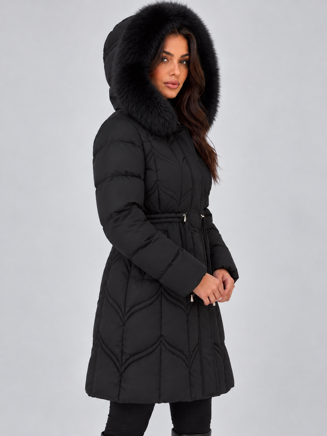 Side view of the black hooded puffer coat with the hood raised, showing the long silhouette, quilted construction, cinched waist, and fur trim.