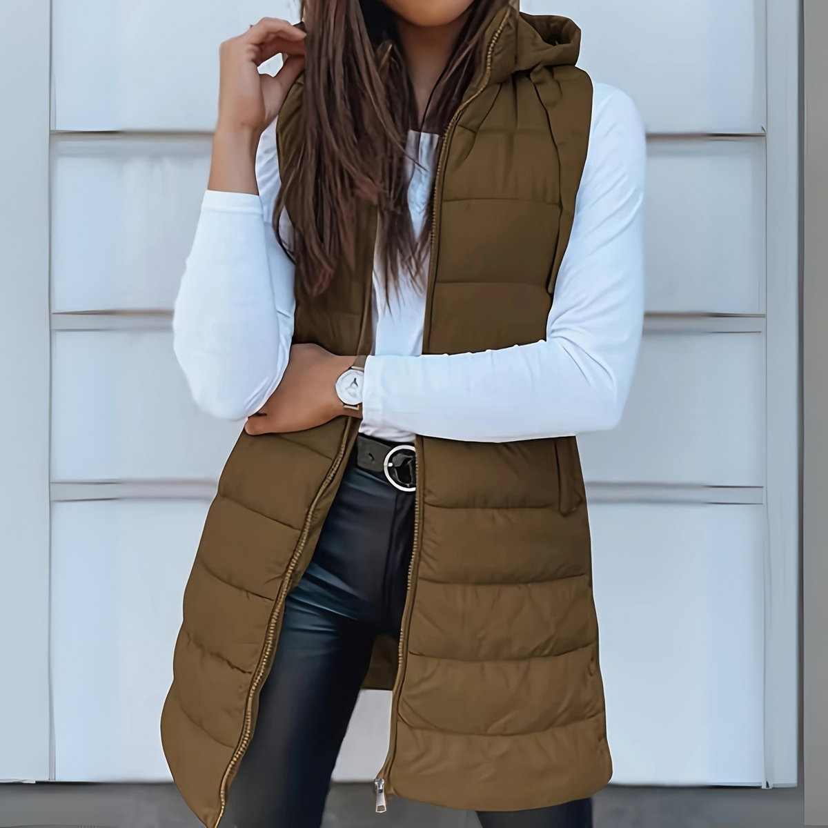 Brown long hooded puffer vest layered over a white top, shown partially zipped with its extended length and horizontal quilting visible.