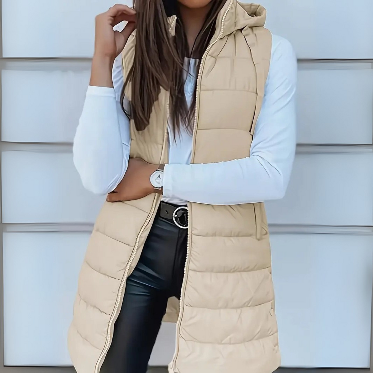 Light beige hooded puffer vest displayed over a white long-sleeve shirt, with the front zipper open and the long quilted silhouette visible.