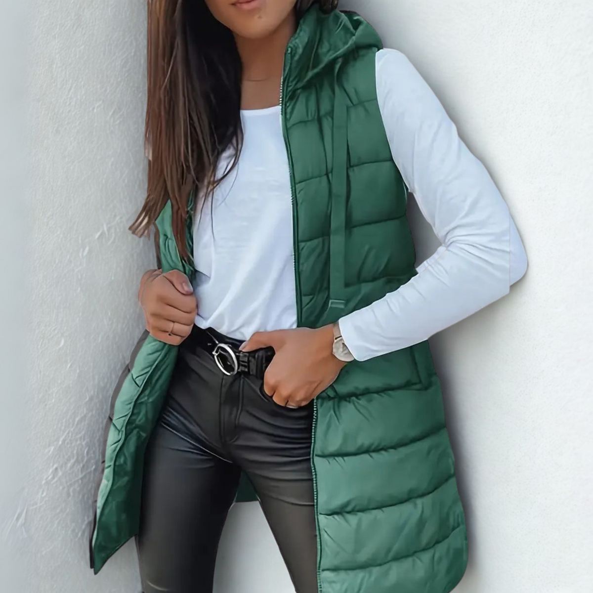Dark green long hooded puffer vest worn open over a white long-sleeve top, showing the streamlined quilted front and extended length.