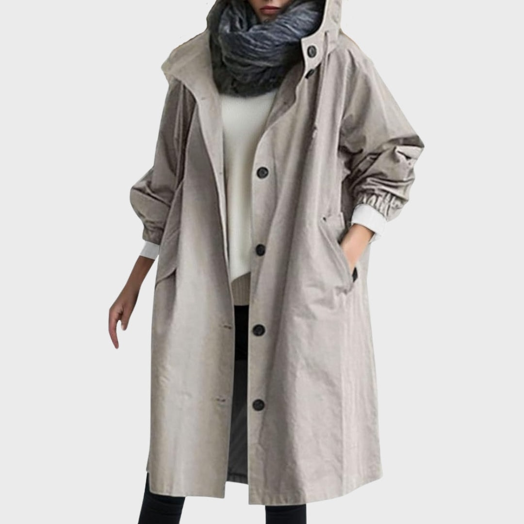Front view of a long light taupe hooded rain coat with button closure, side pockets, and elasticated cuffs over layered clothing.