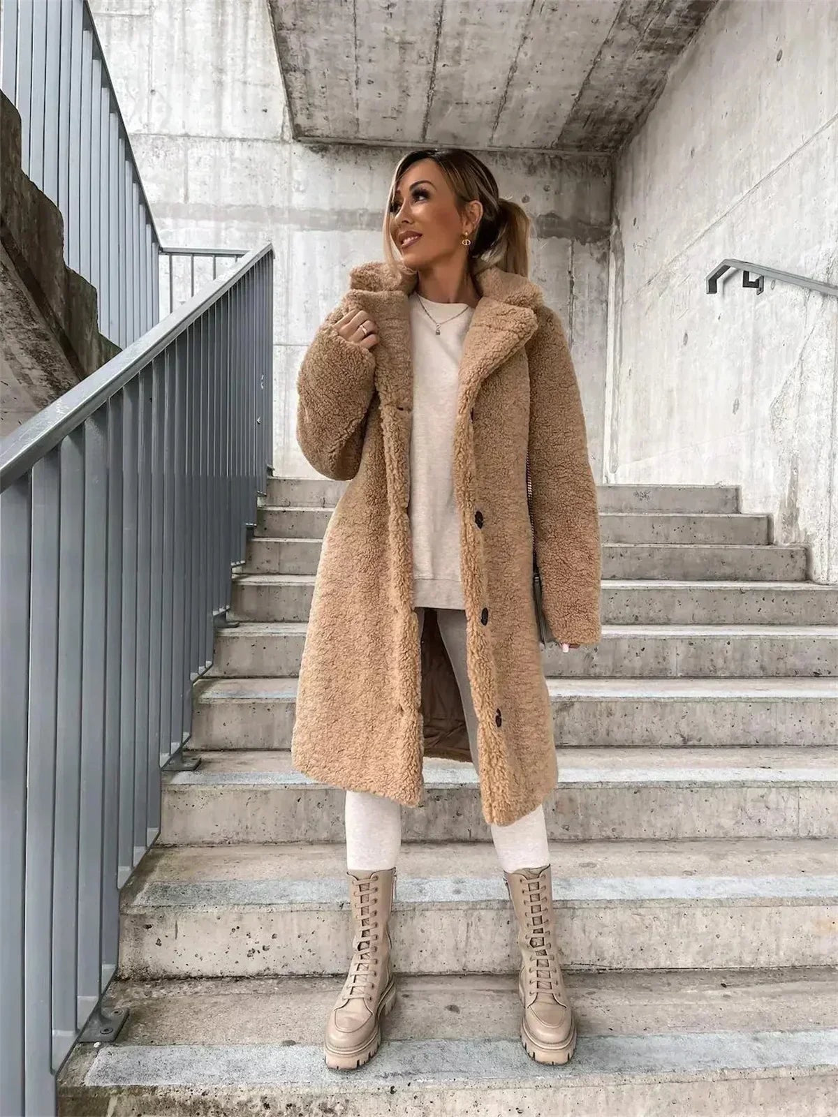 Full-length view of the tan teddy coat on stairs, showing its streamlined below-the-knee shape, notched collar, and button-front closure.