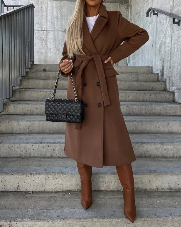 Brown longline double-breasted coat with a tied waist belt, notched lapels, and front flap pockets, worn with a dress and knee-high boots.
