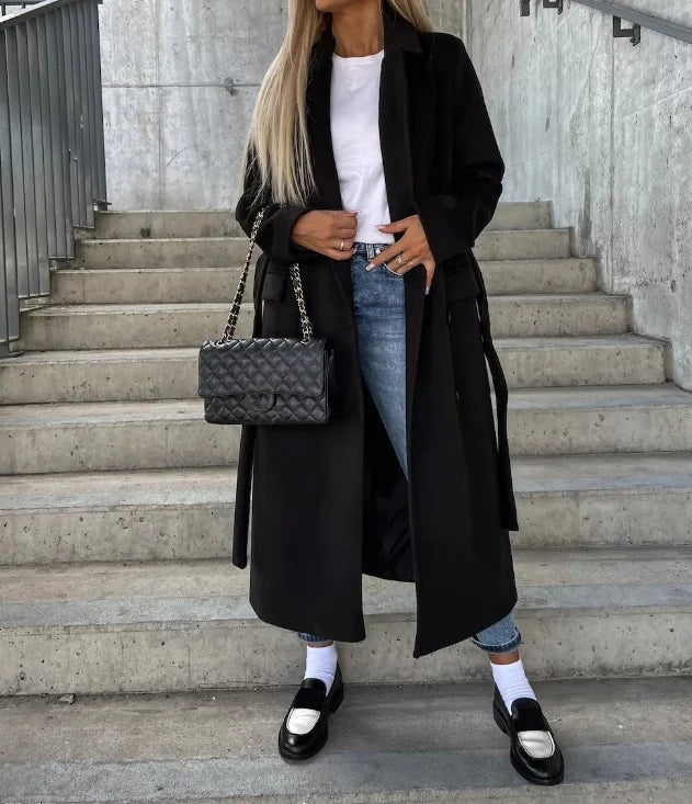 Black longline coat with notched lapels and side pockets, worn open over a white top and jeans.