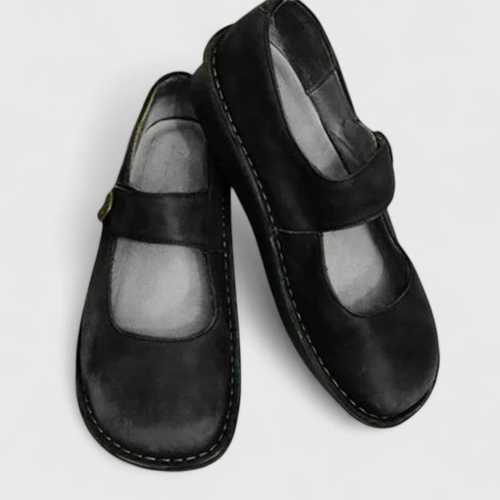 Black Mary Jane flats shown diagonally, with one shoe overlapping the other and the adjustable strap design visible across each instep.