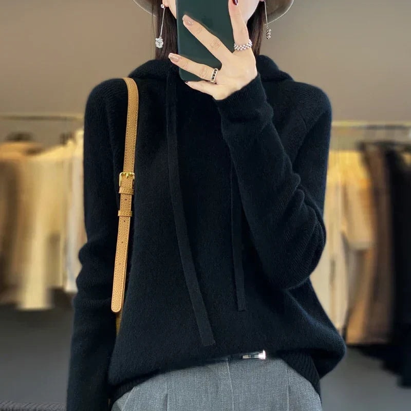 Black oversized knit hoodie with long drawstrings and ribbed cuffs, worn with gray trousers and a shoulder bag.