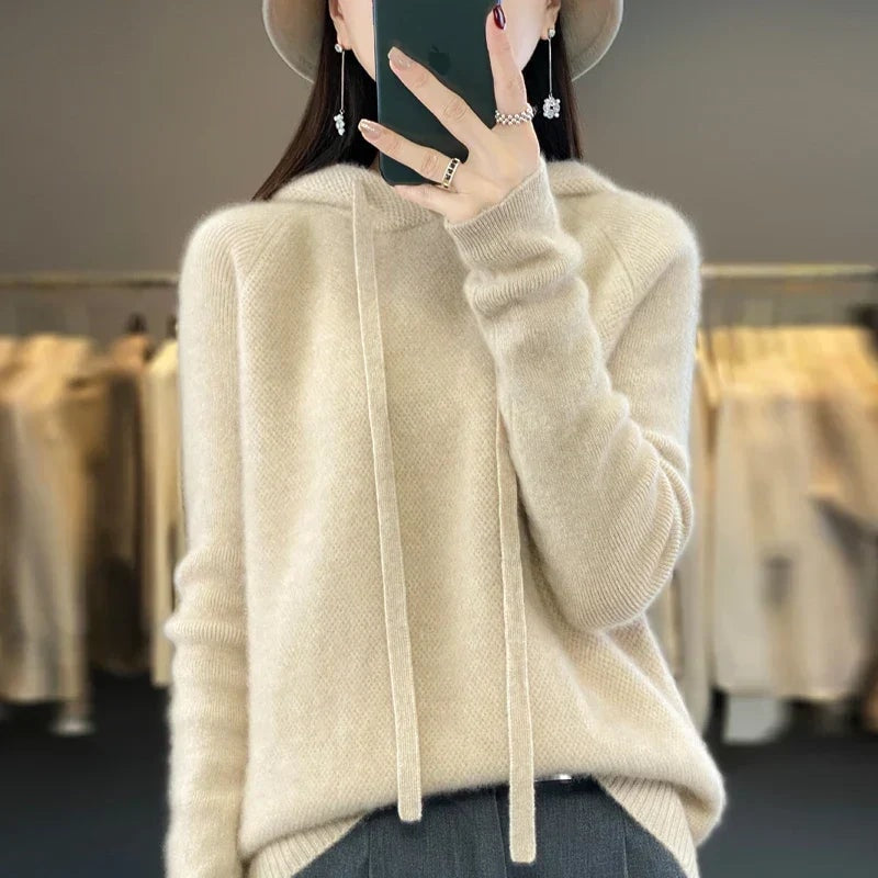 Cream oversized knit pullover hoodie with a roomy fit, long drawstrings, and long sleeves over dark trousers.