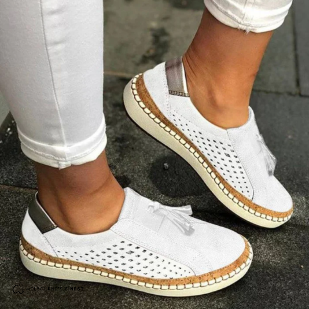 White perforated slip-on sneakers featuring front tassels, metallic-look heel accents, and contrasting stitched soles.