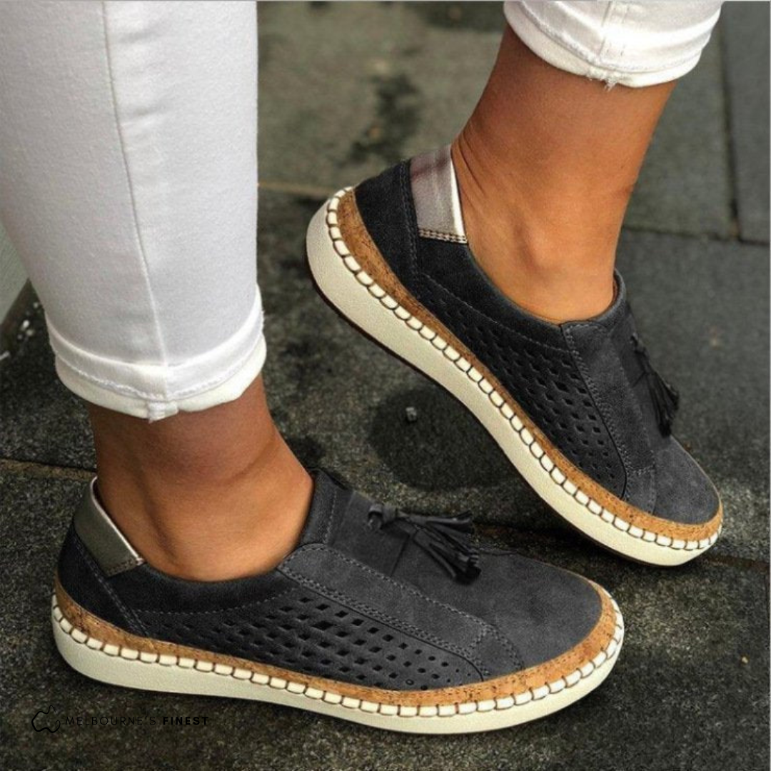 Black perforated slip-on sneakers with front tassel details, metallic-look heel accents, and contrasting stitched soles.