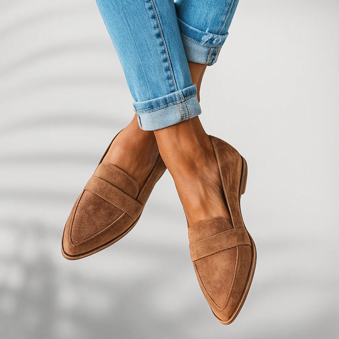 Taupe pointed toe slip-on loafers shown suspended in a crossed-leg pose, emphasizing their sleek profile and layered detailing.