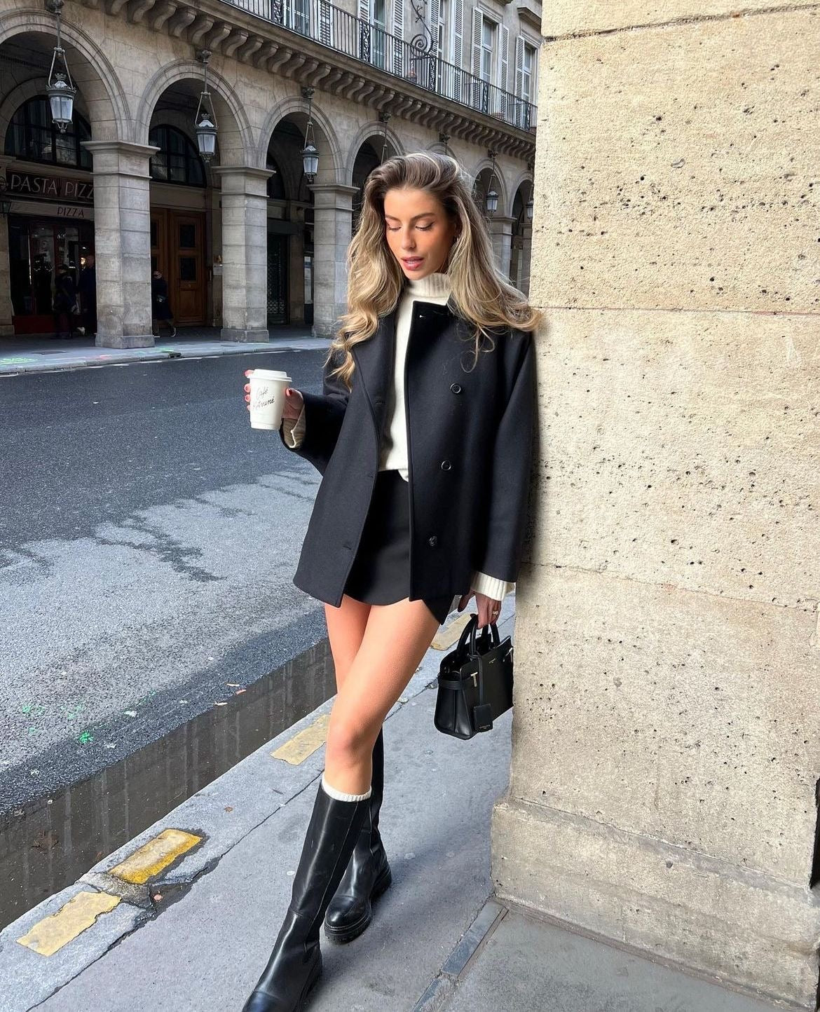 Black short coat worn closed over a light top, styled with shorts and tall boots for a polished city look.