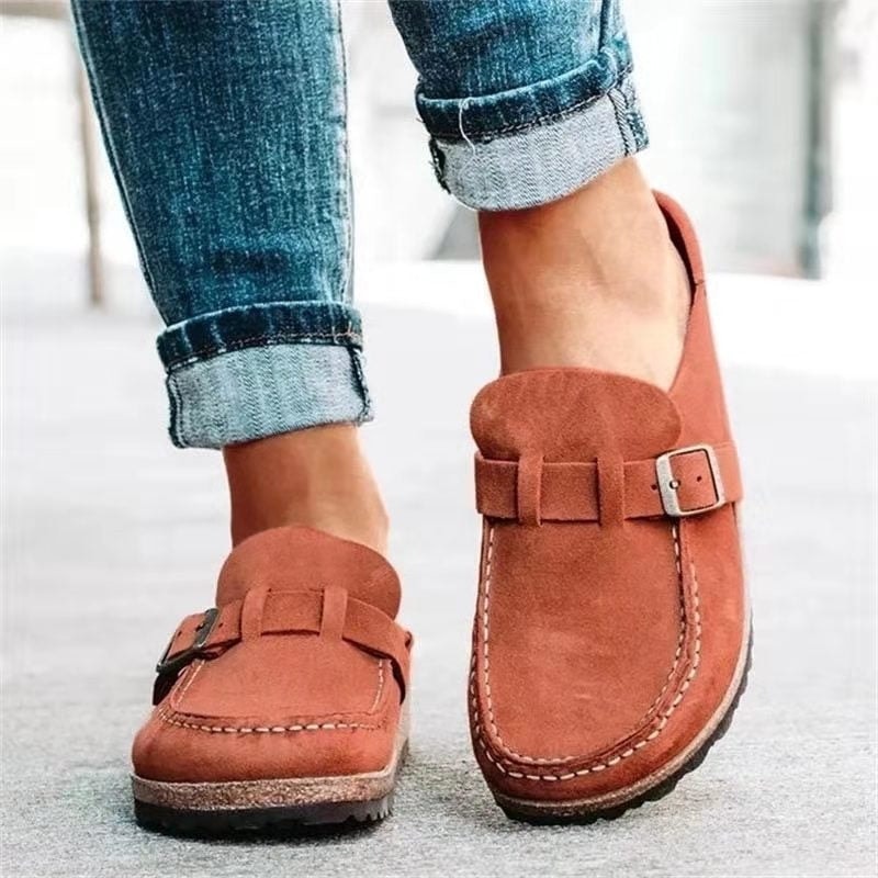 Terracotta slip-on flats with rounded toes, adjustable buckle straps, contrast stitching, and textured low-profile soles shown with cuffed jeans.