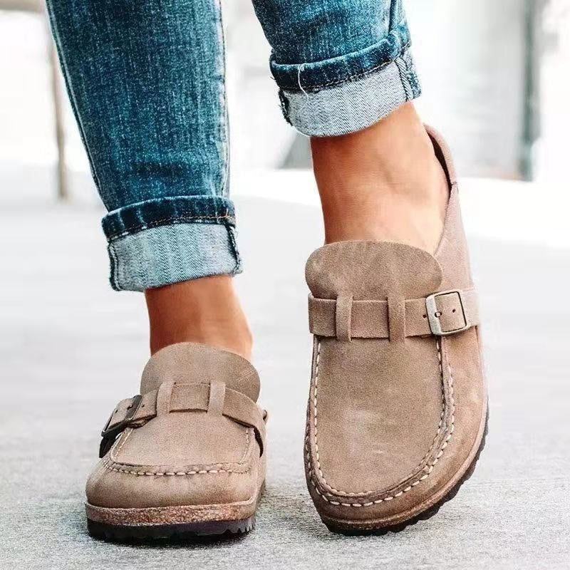 Neutral beige slip-on flats shown from the front and angled side, with rounded toes, top buckle straps, and defined contrast stitching.