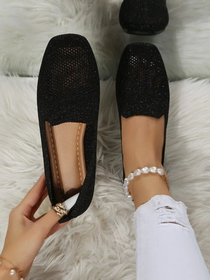 Black open-knit slip-on flats viewed from above, showing their square toes and contrasting foot openings.