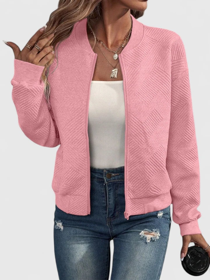 Pink textured geometric-pattern bomber jacket with a round neckline, zip front, long sleeves, and waist-length fit, styled with a white top and distressed jeans.