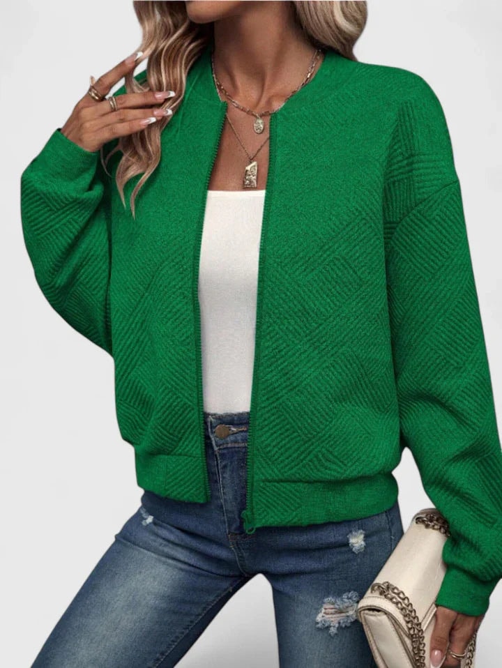 Green waist-length bomber jacket with a raised geometric texture, round neckline, zip closure, and long sleeves, paired with jeans and a light handbag.