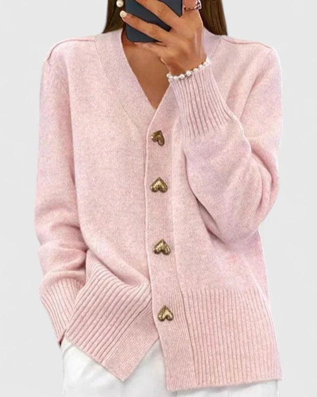 Soft pink V-neck cardigan with gold heart buttons, long sleeves, and ribbed cuffs and hem, worn with white trousers.