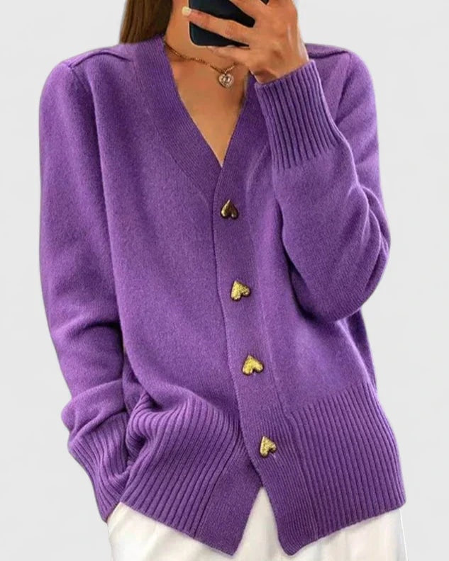 Purple V-neck cardigan with four heart-shaped buttons and pronounced ribbed cuffs and hem, styled over white trousers.